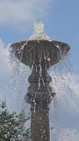 This media file captures a close-up, low-angle view of a decorative fountain on a bright, sunny day. The fountain, constructed of a dark, textured material resembling stone or concrete, features a tiered design with water cascading over the edges of the uppermost bowl. A central spout sends a spray of water upwards, creating a dynamic and refreshing visual. The background consists of a blue sky dotted with white, fluffy clouds, suggesting fair weather. To the left, the blurred foliage of trees hints at an outdoor, possibly park-like or garden setting. The overall impression is one of tranquility and natural beauty, with the sound of the flowing water likely contributing to a serene atmosphere. The footage appears to be captured in Peachtree Corners, United States of America. No visible text is present in the media.