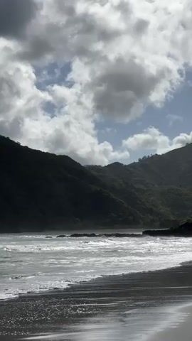 The video depicts a landscape featuring a beach, ocean, and mountains under a partly cloudy sky. The beach appears to have dark sand, possibly black, with ocean waves washing ashore. Lush green mountains form the backdrop, partially obscured by cloud cover. The sky is a mix of white clouds and patches of blue. No people or animals are visible in the frame. The scene is calm, with gentle waves and diffused sunlight.