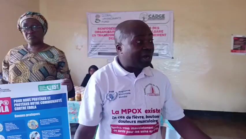 A man in a white polo shirt with logos on it is speaking to the camera in an indoor setting. He is gesturing with his right hand. Next to him on his left, a woman in a headscarf and patterned dress is holding up a poster that says "STOP EBOLA." The poster also has text in French and images related to preventing Ebola. In the background, there is a banner with text in French and logos. Another woman is partially visible in the background. The environment appears to be a room with plain walls.