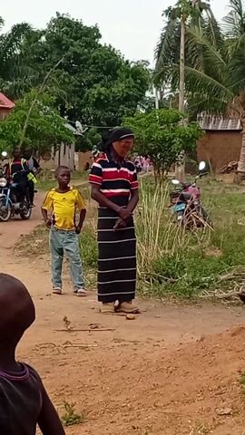 The video shows a woman and a young boy standing on a dirt road in a rural area. The woman is wearing a striped shirt and a long, striped skirt, and she has a head covering. The boy is wearing a yellow shirt and blue jeans. They both look towards the right of the frame. Behind them is a motorbike parked in some grass and some buildings. Another person is standing in the background to the left. The setting is somewhat overgrown. Someone is speaking in the background.