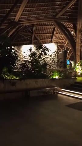 The video captures a nighttime scene of an open-air structure with a distinctive bamboo-like roof. The architecture features curved beams that create an arched effect. The area appears to be an entrance or walkway, with stone walls and lush green plants illuminated by warm, ambient lighting. Steps with integrated lights lead upwards. The ground is paved with a dark material. The overall atmosphere is dimly lit and tranquil, suggesting a resort or an upscale establishment. The camera is moving, providing a sweeping view of the location.