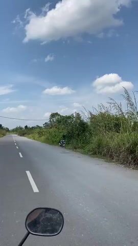 The video shows a first-person perspective of riding on a road. The road is paved with a white line marking the lanes. On the right side of the road, there is a ditch filled with lush green vegetation. A parked motorcycle is visible in the ditch. The sky is a clear blue with scattered white clouds. The overall impression is a scenic drive through a rural or natural landscape.