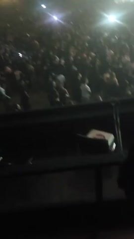 The video shows a large concert crowd in a dark venue lit by spotlights. The camera is positioned at a high angle, looking down onto the audience. Many individuals are visible, closely packed together, some holding up their phones, likely recording the performance. The lighting changes from white to red, and back to white, illuminating the crowd and the stage. In the distance, a performer can be seen on stage, obscured by the bright lights. The overall atmosphere appears energetic and lively.