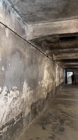 This video shows the interior of a dimly lit, austere room with concrete walls and a low, exposed concrete ceiling. The walls are stained and appear weathered, with patches of peeling paint. Several bare light bulbs are suspended from the ceiling, casting a stark illumination. The floor is also concrete and shows signs of wear.

Two individuals are visible in the mid-ground, walking away from the camera. One person is wearing a dark t-shirt and shorts, while the other is wearing a light-colored t-shirt and pants. They appear to be visitors to the location. In the background, there is a doorway with a barred gate, and further down the hallway, another person can be seen.

The overall atmosphere of the scene is somber and stark, suggesting a place with a significant historical context. The environment is characteristic of a former detention facility or prison, given its stark, utilitarian design and the evident signs of age and disrepair. The location is identified as Oświęcim, Poland, which is widely known as the site of the Auschwitz-Birkenau concentration and extermination camp. The video captures the interior of a structure within this historical complex. No text is visible in the video.