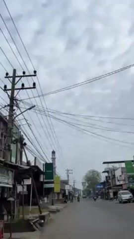 A vertical shot captures a street view on a cloudy day. The street, lined with buildings and various storefronts, stretches out into the distance. An OPPO sign is visible on one of the buildings, along with a smaller Realme advertisement. Overhead, numerous power lines intersect and weave across the sky. In the background, a mosque's minaret is discernible. Vehicles, including motorcycles and cars, traverse the street. Additionally, a couple wearing traditional Muslim attire walks along the sidewalk. The sky overhead is overcast, with a uniform layer of clouds.