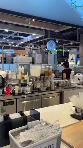 The video shows a busy beverage counter in what appears to be a mall or food court. In the foreground, there is a white counter with a basket of plastic cups and straws. To the left, a black organizer holds various items. Behind the counter, stainless steel equipment is visible, including a beverage dispenser with multiple taps, bottles of syrups and liquids, and cups. A person wearing a red shirt and black apron is working behind the counter, preparing drinks. The lighting is bright, and there are signs and logos in the background, including a circular sign with text that appears to be in Thai. The overall atmosphere suggests a commercial establishment focused on serving drinks.