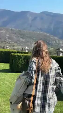 The video shows a person walking on a green lawn. The person has long brown hair, wears a plaid shirt over a white top, and white pants. They are carrying a tan trench coat over their arm and have a brown purse slung across their shoulder. A small dog with black and white markings runs around the person. In the background, there are neatly trimmed green hedges, white decorative orbs, and distant mountains under a clear blue sky. The scene appears to be a formal garden or a landscaped area with well-maintained grass. The camera follows the person as they walk across the lawn.