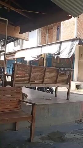 The camera pans right from an interior space to a street view. In the foreground, there are two wooden benches. The street is unpaved and has some trash on it. Two motorcycles are parked on the side of the street. In the background, there are several brick and concrete buildings with metal roofs.
