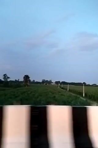 The video shows a road through a field under a cloudy sky. In the foreground, a blurred barrier with black and white stripes is visible. On the road, a vehicle with its headlights on is approaching. To the left of the road, there is a water tower and some trees. The field on both sides of the road appears to be covered in tall green crops. A bicycle is mounted on a pole on the side of the road. The scene suggests travel through a rural area, possibly during dusk.