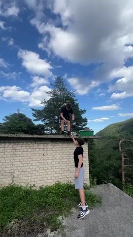 Two young men are on a brick structure overlooking a lush green valley under a bright blue sky with scattered white clouds. The man in the foreground is wearing a black t-shirt and grey shorts, standing with his hands at his sides and looking at the camera. The man in the background is wearing a black t-shirt and black shorts and is standing on the edge of the structure, looking down at his phone. A tall evergreen tree is behind him. The environment is natural and mountainous, with dense vegetation covering the slopes of the valley.