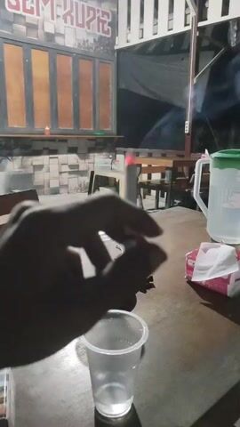 A person's hand is visible, holding a cigarette that is actively being smoked, with smoke rising from it. The cigarette is positioned over a clear plastic cup on a wooden table. In the background, there are tables and chairs, suggesting an outdoor seating area, possibly a cafe or restaurant. A large green-lidded jug and a packet of tissues are also on the table. The lighting indicates it is nighttime. A sign with text "SEM-KUPIE" is visible on a wall in the background.