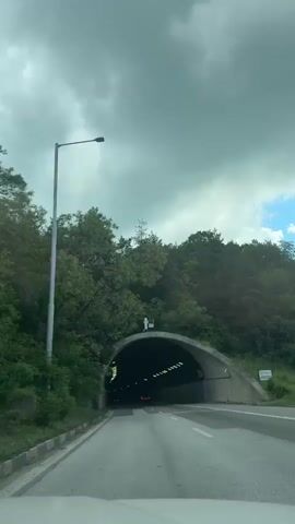 The video shows a car's perspective entering a tunnel. The tunnel entrance is arched and made of concrete. Lush green trees and foliage surround the tunnel entrance. The sky is overcast with dark clouds. Inside the tunnel, lights line the ceiling at regular intervals, illuminating the road. The road is paved and has white lane markings. There are faint red lights visible in the distance within the tunnel, suggesting other vehicles. The camera appears to be mounted inside the vehicle, looking forward.