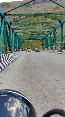 This media file captures a scenic view of a bridge in Teling, India, likely from the perspective of a motorcycle. The bridge is a prominent green metal structure with a latticework design, spanning over a road. The road itself is asphalt and appears to be a two-lane highway. On either side of the road, there are black and white striped barriers, adding a visual element to the scene.

In the distance, a white van and a dark-colored car are visible on the bridge, suggesting traffic is present. Along the sides of the bridge, there are prayer flags, a common sight in mountainous and spiritual regions of India, indicating a cultural context. Several people can be seen standing on the side of the bridge, possibly observing the surroundings or waiting.

The background of the scene is dominated by a lush green mountainside, characteristic of hilly terrain. The sky appears to be overcast or hazy, suggesting a cloudy or possibly rainy weather condition, but the overall lighting doesn't indicate a specific time of day. The presence of mountains and prayer flags strongly suggests a location in a mountainous region of India, consistent with the provided location context of Teling. The overall mood of the media file is one of travel and exploration, with a sense of natural beauty and cultural significance. No discernible text is visible in the media file.