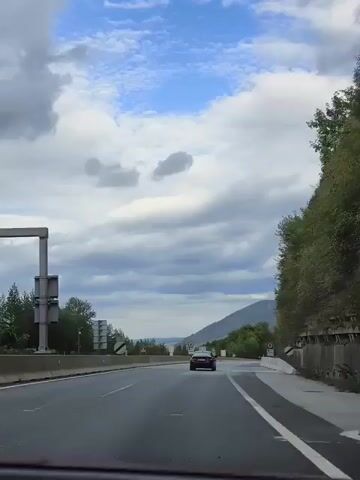 The video shows a car driving on a highway under a cloudy sky. The highway has multiple lanes and a concrete barrier on the right side. There are trees and vegetation on the right side of the highway, and a hillside in the distance. The car is red and is traveling in the right lane. The sky is filled with white and gray clouds, with some patches of blue sky visible. The overall environment appears to be a rural or semi-rural area. The footage appears to be captured from another vehicle moving in the same direction.