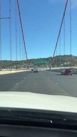 A point-of-view shot from inside a vehicle, showing the roadway of a bridge. Green overhead signs indicate directions to Oakland, San Francisco, Cummings Skyway, Crockett, and Pomona Street. Traffic moves along the bridge, with several cars and a white van visible. The sky is clear and blue, and a hilly, tree-covered landscape borders the bridge. The perspective suggests movement forward at a moderate speed.