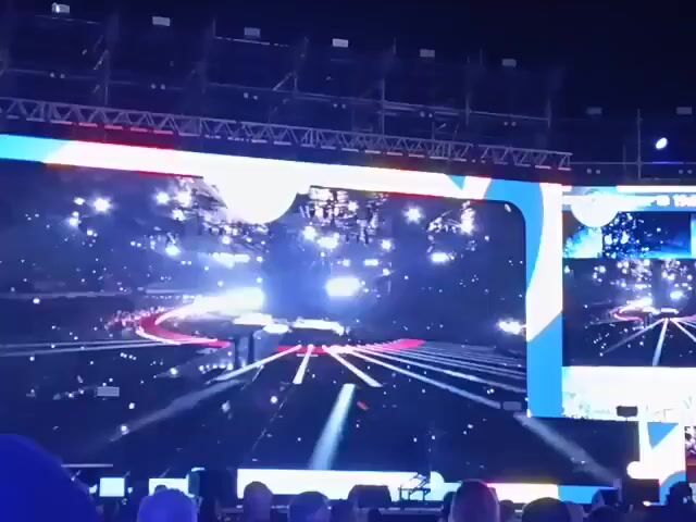 Here is a description of the video:

The video shows a large outdoor concert stage with two large LED screens. The screens display the word "POLAND" in red and a graphic of a futuristic-looking stage with white lines and red accents. The text below "POLAND" lists names, possibly of performers or crew members. There is a crowd of people watching the stage. The video pans to show the crowd more clearly. The night sky is visible above the stage, and some city lights can be seen in the background. The atmosphere appears festive.