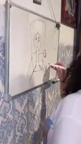 The video shows a person standing in front of a whiteboard, drawing a figure with a marker. The person is wearing a white shirt and has curly hair. The whiteboard is mounted on a wall with a patterned wallpaper. There is also a picture frame hanging on the wall next to the whiteboard. The figure drawn on the whiteboard has a large head, a simple face, and a body with arms and legs. The figure is also holding something in one hand. The person is focused on drawing and does not interact with anyone else in the video.