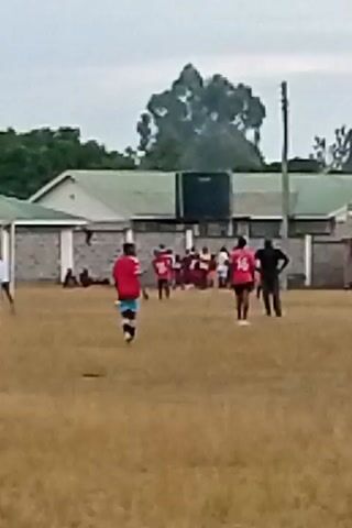 The video shows a group of people on a grassy field, likely a sports field or park. Most of the people are wearing red shirts, and some are wearing other colored clothing like blue, white, purple, yellow and black. One person wearing a red shirt with the number 16 on the back is walking away from the group. Behind the people, there is a building with a light green roof. There are also trees in the background. The sky is overcast. The video appears to be taken from a distance, and the image is somewhat blurry.