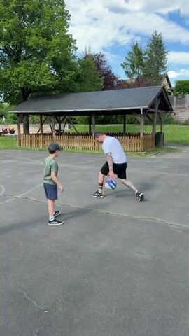 A man and a boy are on an outdoor basketball court. The boy is wearing a green t-shirt, dark shorts, a baseball cap, and sneakers. The man is wearing a white t-shirt, dark shorts, and sneakers. He has tattoos on his arms and legs. He is holding a ball that is red, white, and blue. The man is dribbling the ball and then kicking it to the boy. Another boy walks into the frame. In the background is a covered picnic area with a wooden fence. There is a green tree in the background and a building with a pitched roof. The sky is blue with scattered clouds. The basketball court is asphalt with white lines.