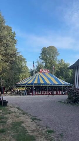 The video shows a large, striped circus tent with yellow and blue sections, pitched in an outdoor area. The tent has "CIRCUS BAR" written in red letters along the bottom of the structure. There are people inside and around the tent, some appearing to be customers and others possibly staff. The tent is supported by poles and ropes, and two French flags are visible at the top. The setting appears to be a park or wooded area, with trees surrounding the tent and a gravel path in the foreground. The sky is blue with some clouds, and the lighting suggests it is daytime.