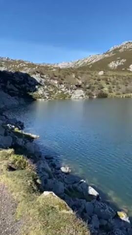 The video shows a still shot of a blue lake surrounded by rocky, hilly terrain. The sky is clear and blue. The camera slowly pans from left to right, revealing the lake and the surrounding landscape. A voice can be heard discussing Christina Aguilera.