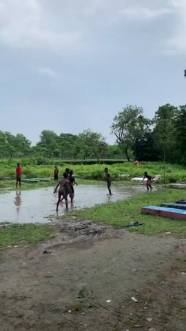 A group of seven young boys are playing in a flooded, muddy field on a cloudy day. They are all shirtless and appear to be playing a game involving a ball. The field is surrounded by lush green trees and vegetation. The ground is wet and muddy, with several puddles of water scattered throughout. In the background, there are some buildings or structures. The sky is overcast with gray clouds. The boys are actively engaged in their game, kicking and chasing the ball through the water. One boy in the foreground is particularly animated, splashing water as he moves. Another boy in the background is wearing a red shirt. The overall scene depicts a lively and energetic moment of childhood play in a rural setting.