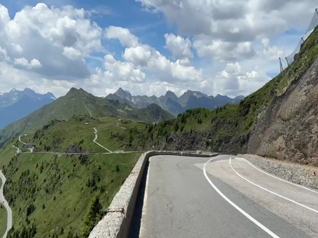 The video shows a winding mountain road with a stone retaining wall on the left side. The road curves to the right, and the camera pans across a vast mountain landscape under a partly cloudy blue sky. In the distance, multiple mountain ranges are visible, some with snow-capped peaks. The slopes of the foreground mountains are covered in green grass and scattered evergreen trees. A few small buildings are visible on the mountainside in the mid-ground. The road has white lane markings. The video captures a panoramic view of a scenic mountain pass.