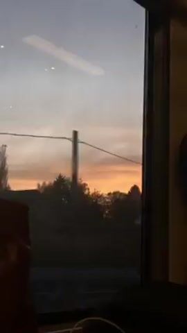 From the vantage point of a train passenger, the camera peers through a window at dusk, with a skyline painted in shades of orange and violet. In the foreground, glimpses of the train seat's headrest and a cable indicate an interior setting. The window's reflection reveals interior lights. Across the tracks, a silhouetted house is briefly visible, followed by trees against the fiery horizon.
