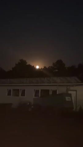 The video shows a nighttime scene with a bright full moon partially obscured by dark trees. In the foreground, there is a long, low, white building with several windows. In front of the building, partially silhouetted, are a few objects including what appears to be a vehicle or trailer. The moon is positioned centrally in the sky, above the tree line. The overall ambiance is dark and serene.