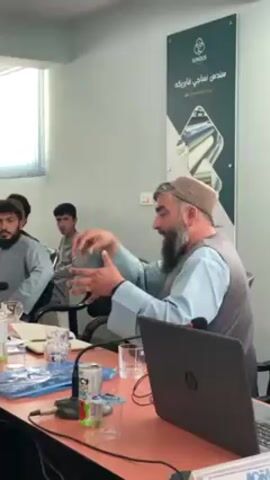 An indoor scene depicts a meeting or discussion in what appears to be an office or conference room. In the foreground, a bearded man, dressed in a light blue traditional shirt, a dark vest, and a light-colored cap, is speaking animatedly, gesturing with both hands. He is seated at a wooden table with a black HP laptop open in front of him, flanked by microphones.

Around the table, several other men are present, listening attentively. They are also dressed in traditional attire, with some sporting beards and caps. One younger man in the background is clean-shaven.

The table is cluttered with typical meeting items: clear glasses, a water bottle, a can of Coke Energy, and a blue folder containing documents. The room itself has light-colored walls, and natural light streams in from a window visible on the left, suggesting it is daytime.

On the wall behind the speaker, a large poster is displayed. It features the English word "SUNDUS" and text in Dari/Pashto, which reads "سندس نساجي فابريكه" (Sundos Nesaji Fabric), indicating a business or industrial context. The overall atmosphere suggests a focused and engaged professional gathering.