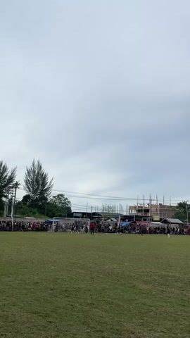 A wide shot captures a soccer game on a grassy field under a cloudy sky. A crowd of spectators lines the perimeter of the field, with some standing and others sitting behind temporary structures and fences. In the background, buildings under construction are visible, along with trees and utility poles. Several players are on the field, and a soccer goal is positioned on the left side of the frame. The game appears to be in progress.