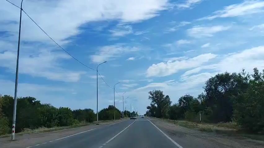 The video shows a POV shot from a vehicle traveling down a paved road. The sky is blue with scattered white clouds. There are streetlights lining the right side of the road. Dense green foliage lines both sides of the road. Several cars are visible on the road ahead. The road appears to be in a rural or semi-rural area.