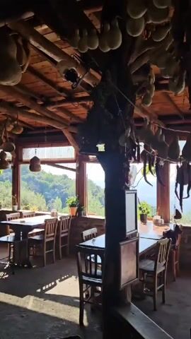 The video showcases the interior of a rustic restaurant, with a wooden ceiling adorned with numerous hanging gourds and other dried items. The camera pans slowly from right to left, revealing several wooden tables and chairs set up for dining. Large windows along the back wall offer a view of a lush green landscape outside. A few patrons are visible seated at tables in the distance. Towards the left side of the frame, there's a counter area with various kitchen items and decor. The lighting is natural, casting shadows across the floor.