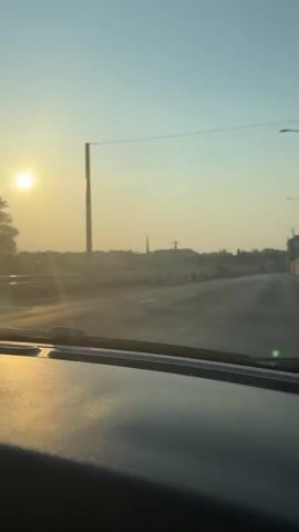 The video shows a view from inside a vehicle, looking out through the windshield. The sun is low in the sky, casting a bright glow and lens flares across the scene. The road ahead is wet, with reflections of the sunlight visible on the surface. Construction barriers, marked with red and white stripes, are present along the side of the road. In the distance, there are buildings, trees, and a tall spire, likely belonging to a church. Utility poles and wires are also visible against the bright sky. The overall impression is of a journey during sunrise or sunset.