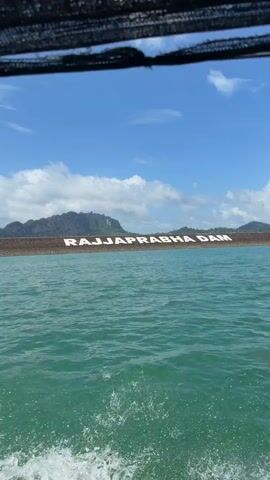 The video shows a wide shot of a large body of water, likely a lake or reservoir, with a dam visible in the background. The dam has large, white letters spelling out "RAJRAPRABHA DAM." The water is a turquoise green and appears choppy, with white foam from a boat's wake at the bottom of the frame. The sky is blue with scattered white clouds. The perspective suggests the video is being filmed from a moving boat. No people are clearly visible in the frame.
