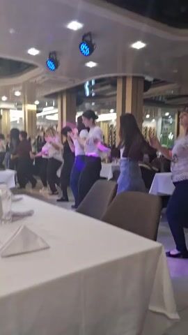 In an indoor setting, a group of roughly a dozen women of varied ages, mostly dressed in casual attire, are engaged in a line dance in a restaurant or banquet hall. A man approaches the group. Several tables set for diners are visible. The lighting is provided by overhead fixtures as well as colorful stage lighting that changes color. The floor appears to be tiled.