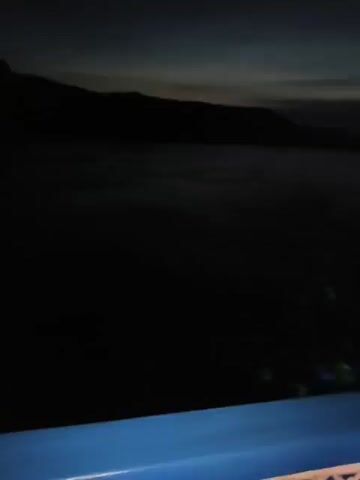A night-time aerial video begins with a view of a dark landscape. The camera pans slowly across a body of water, its surface rippled by a gentle breeze. In the distance, the silhouette of a mountain range is visible against a dark sky. The video transitions to a view of the horizon, where a faint glow indicates the approaching dawn. The sky gradually lightens, revealing streaks of color as the sun begins to rise. The camera continues to pan, showing more of the mountain range and the vast expanse of water below. The video concludes with a close-up of the water's surface, reflecting the soft light of the rising sun.