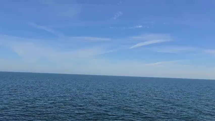 The video begins with a wide shot of the ocean and sky. As the camera zooms out, a tall ship with three masts and green sails becomes visible in the distance, sailing across the calm blue water under a partly cloudy sky. The ship is the sole object in the frame apart from the sea and sky.