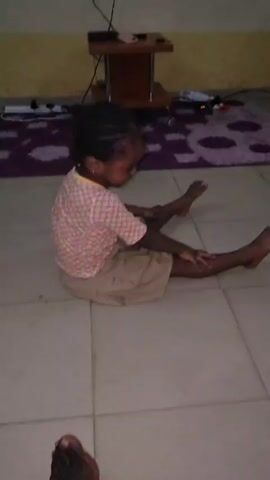 A young, dark-skinned child with braided hair sits on a white tiled floor, then gets up. The child wears a patterned shirt and tan shorts. A purple and white rug is visible in the background, along with a small brown table with a phone on it and various electronic devices and cords. Another person's bare foot is visible in the lower left of the frame. The walls are yellow.
