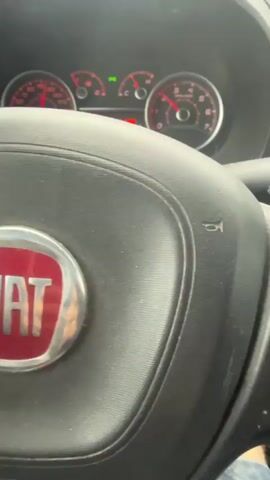 The video shows the interior of a car, focusing on the dashboard and steering wheel. A hand is visible giving a thumbs-up gesture. The car is in motion on a wet road, as indicated by the windshield wipers actively clearing rain. The dashboard displays the car's speed, fuel consumption, and other indicators, suggesting the car is being driven in rainy weather conditions. The overall scene depicts a person driving in inclement weather.
