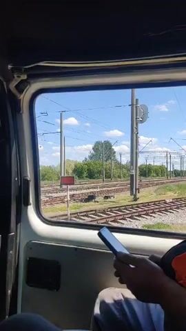 The video captures a view from inside a vehicle, looking out a side window at a railway environment in Shelestove, Ukraine. The vehicle appears to be moving slowly alongside multiple parallel train tracks on a sunny day with a bright blue sky and scattered white clouds. Overhead power lines for the railway system are visible, supported by numerous utility poles. Green trees and bushes line the horizon.

In the foreground, a person's hands are visible, holding a black smartphone, seemingly engaged with it. As the vehicle proceeds, it passes a set of white utility boxes or equipment. Subsequently, a white and yellow sign comes into view, displaying the Cyrillic word "ПОСТ" (Post) at the top and "ОБЪЕЗД" (Detour/Bypass) written vertically below it. Shortly after, a small, light-colored building with a green roof and blue-framed windows is seen before the view returns to the railway tracks and surrounding vegetation.