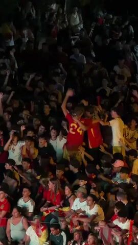 The video shows a large crowd of people, mostly young adults and teenagers, watching something off-camera. Many are wearing red and yellow, possibly indicating support for a sports team or national flag. The atmosphere appears festive, with some individuals raising their arms and cheering. The lighting is dim, suggesting it is evening or night. Some people are holding small flags. The perspective is from behind the crowd, looking down.