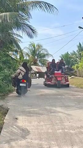 A motorcycle with two people on it speeds down a dirt road lined with palm trees and vegetation. A red, three-wheeled vehicle with two people riding in the front passes them. The sky is blue and clear.