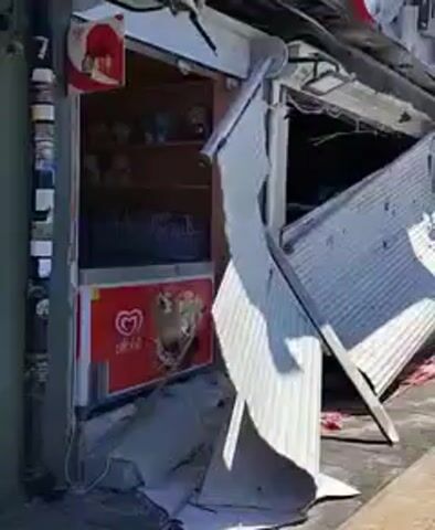 The video shows a kiosk that has sustained heavy damage. The corrugated metal roll-up door is buckled and partially detached, revealing the interior. Inside, shelves are visible stocked with various items. A red cooler with the “Algida” ice cream logo is also visible inside the kiosk. Outside, debris is scattered around the base of the structure, and the kiosk appears to be located on a sidewalk. In the background, other buildings and traffic can be seen, indicating an urban setting.