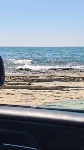 The video captures a view from inside a vehicle looking out at a rocky coastline. In the foreground, the dark interior of a car, possibly a door panel and a side mirror, is visible. The mirror reflects a distorted image of the sky and the car's interior. Beyond the car's edge, a textured, light-colored rocky shore is prominent, with horizontal layers and grooves. The sea, a vibrant blue, meets the shore with gentle waves breaking on the rocks, creating white foam. The horizon is clearly defined between the sea and a clear, pale blue sky. The lighting suggests it is a sunny day. No people are visible in the frame.