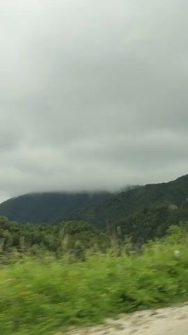 The video shows a view from inside a vehicle, looking out at a cloudy, mountainous landscape. Green trees and vegetation are visible in the foreground and on the slopes of the mountains. A large sign is briefly visible on the right side of the frame, showing images of buildings and possibly some symbols. The sky is overcast with heavy gray clouds. The camera appears to be moving, as evidenced by the motion blur in the foreground vegetation.