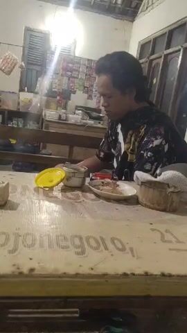 The video shows a person sitting at a table with food. The person has dark, medium-length hair and is wearing a black t-shirt with a colorful, possibly graphic, design. The table is made of wood and has writing on it. On the table, there is a white bowl with a yellow lid, a plate with food, and what appears to be a small woven basket. In the background, there are shelves with various items, including what look like packets of food or condiments. The lighting suggests an indoor setting, possibly a kitchen or dining area, with artificial light sources visible. The overall atmosphere appears casual.