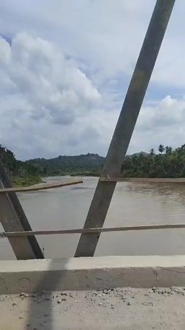 The video shows a view from a bridge overlooking a muddy river. On the left and right sides of the frame, there are large metal beams supporting the bridge structure. In the middle of the frame, a thin pole with the Indonesian flag is visible. The river flows through a lush green landscape with hills in the background. The sky is partly cloudy. The camera pans slightly to the left and right, offering a dynamic perspective of the scene.