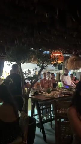 A group of people are gathered at tables in an outdoor dining area. The ceiling is covered in straw or thatched material, and there are hanging lamps. A tree is visible on the left side of the frame. Some people are standing, while others are seated at the tables, eating and drinking. Several individuals appear to be interacting and socializing. The lighting is dim, creating a warm and inviting atmosphere. The overall scene suggests a lively gathering or celebration.