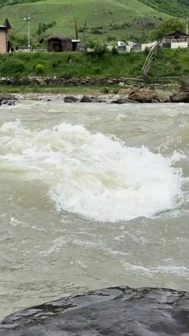 The video shows a river with rushing water. The water is a murky grey color, and there are white foamy waves. In the background is a grassy hill and a few buildings. The video focuses on the river and the sound of the water rushing is audible. There is a large grey rock in the foreground. The river appears to be fast moving and somewhat turbulent.